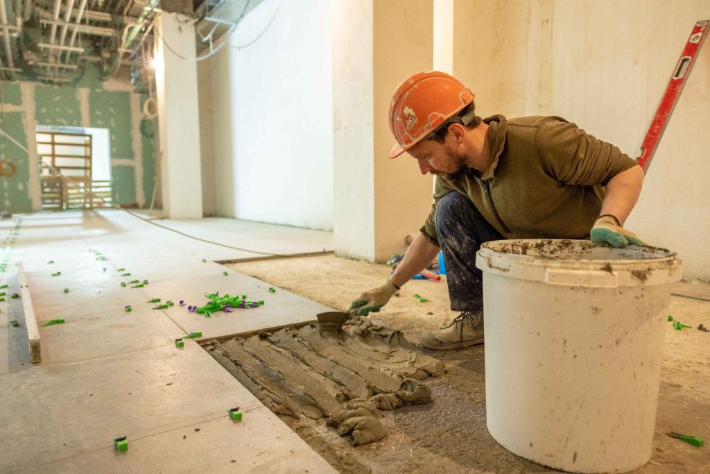 Fliesenleger mit orangem Schutzhelm verlegt Bodenfliesen auf einer Baustelle