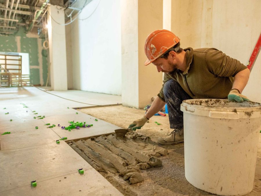 Fliesenleger mit orangem Schutzhelm verlegt Bodenfliesen auf einer Baustelle